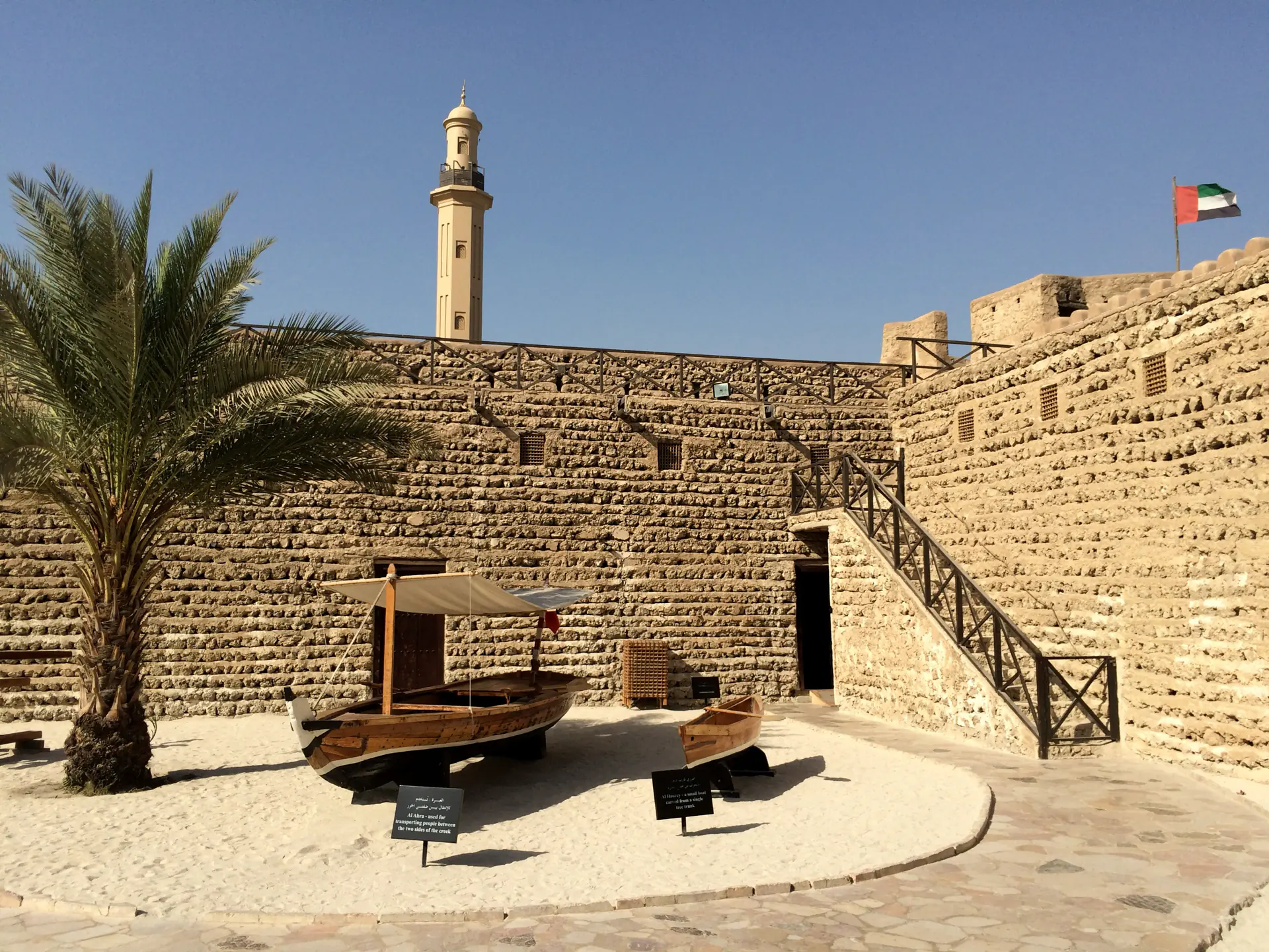 The coral-stone wall of Al Fahidi fort in old Dubai, a palm and old boats beside it.